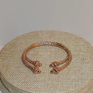 May include: A rose gold-toned open cuff bracelet featuring a textured, woven-style metal band. The two ends of the bracelet terminate in decorative, rounded, knob-like finials with a ribbed pattern, creating a distinct, symmetrical design.