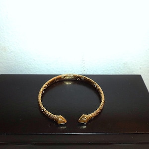 May include: A gold-coloured cuff bracelet with a textured, woven design. The bracelet has pointed end caps. The jewellery is displayed on a dark surface.