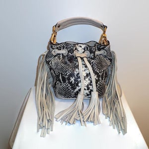 May include: A black and white snakeskin-patterned bucket bag with a white handle and gold-colored hardware. The bag features long, cream-colored fringe and a drawstring closure. A stylish accessory.