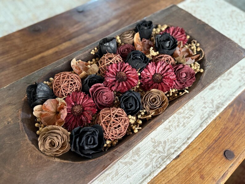 This shows a large coffee table display with a wood dough bowl. The wood is has a dark, moody arrangement of wood flowers and dried botanical berries.