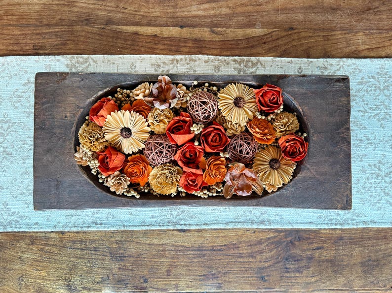 This shows a long wood dough bowl with and arrangement of wood flowers and natural dried botanical berries. The display sits on a coffee table.