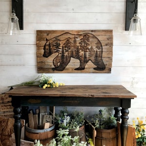 Extra large wall art. Made with reclaimed wood, and features a silhouette of a black bear with a forest scene. Artwork is a wood burning by hand. Wood is a weathered brown. The artwork is dark, burned brown.