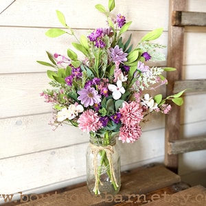 This image shows an artificial bouquet of flowers in a mason jar vase. It features purple daisies, pink mums, and white hydrangeas.