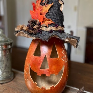 A Large handmade pumpkin witch used for fall decorating. Made from a real dried gourd. The pumpkin, named Hocus Pocus, has a tall burlap crinkled witch hat with a fall leaf arrangement. The pumpkin includes a battery-operated candle.