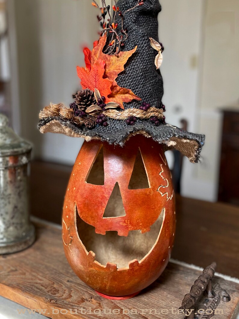 A Large handmade pumpkin witch used for fall decorating. Made from a real dried gourd. The pumpkin, named Hocus Pocus, has a tall burlap crinkled witch hat with a fall leaf arrangement. The pumpkin includes a battery-operated candle.