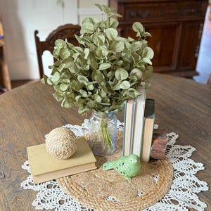 This shows a rustic  table display centerpiece featuring a bouquet of green eucalyptus. The greenery arrangmentsitis in the middle of a round dining room table on topof a crochet doily. Vintage stacked books and a ceramic bird sit near.