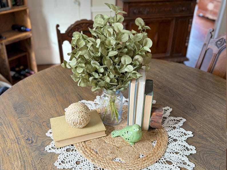 This shows a rustic  table display centerpiece featuring a bouquet of green eucalyptus. The greenery arrangmentsitis in the middle of a round dining room table on topof a crochet doily. Vintage stacked books and a ceramic bird sit near.