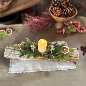 Puede incluir: Un portavelas rústico de tronco de madera con una vela blanca, rodeado de vegetación, rodajas de naranja secas y campanas plateadas. El portavelas está colocado sobre un camino de mesa de lino blanco.