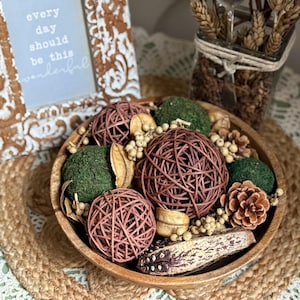 May include: A wooden bowl filled with decorative items, including woven spheres, green moss balls, dried seed pods, and a pine cone. A framed sign with the text "every day should be this wonderful" is in the background.