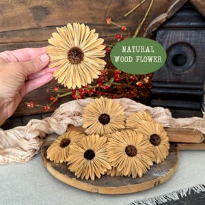 This shows several wood flowers that look like Gerba Daisies. They are laid our on a beige table runner, on top a a vintage style bread board. I am holding one blossom up to show the size and scale of the flower.