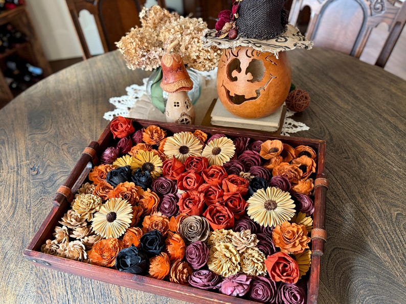 This shows a large wood tray sitting on a dinning room table. The tray is shallow and is holding a large assortments of wood flowers. The flowers are shown in many fall colors such as orange and burgundy. A pumpkin gourd sits in the back.
