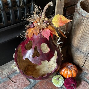 This image shows a fall Halloween decoration. It is a real dried gourd with a carved face.  The gourd is sitting on the fireplace hearth. The face has a ghostly expression. The gourd is stained berry color. It has fall leaves arranged on the top.