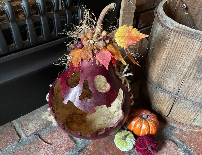 This image shows a fall Halloween decoration. It is a real dried gourd with a carved face.  The gourd is sitting on the fireplace hearth. The face has a ghostly expression. The gourd is stained berry color. It has fall leaves arranged on the top.