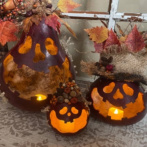 This shows three pumpkins sitting on an entryway table. The pumpkins are made from real gourds. They are stained berry color. Each pumpkin has a ghostly expression carved for a face. The pumpkins are lit up with battery-operated candles.