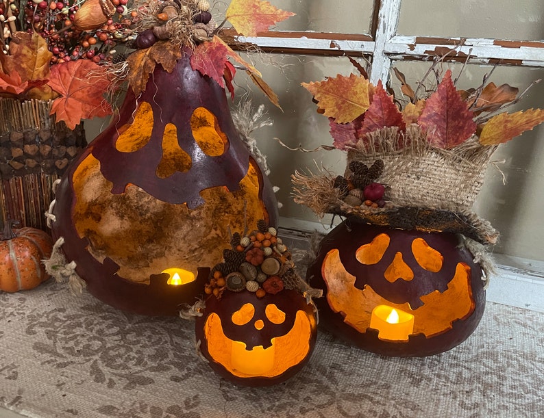 This shows three pumpkins sitting on an entryway table. The pumpkins are made from real gourds. They are stained berry color. Each pumpkin has a ghostly expression carved for a face. The pumpkins are lit up with battery-operated candles.
