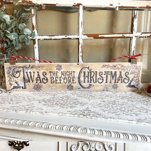 This image shows a handmade wood Christmas sign. It has old-time writing that says Twas the night before Christmas. The decorative writing is wood burned on a plank of reclaimed wood. It has two metal loops with a red and brown rope tied to the top.