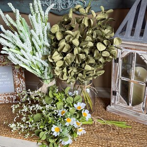 This shows green and white faux flower arrangements on an entryway table. A bouquet of daisies are tied together and lay in front of bouquets of eucalyptus and white lavender. A distressed lantern with a candle sits near.
