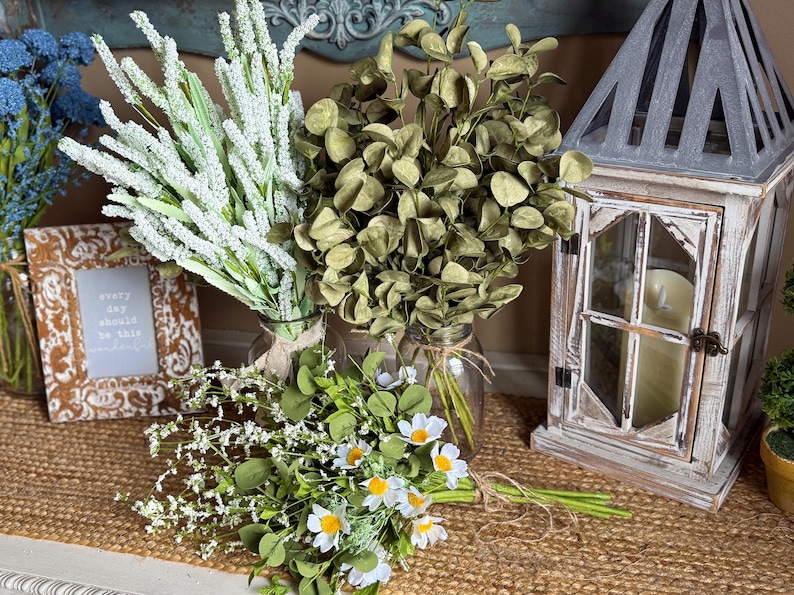 This shows green and white faux flower arrangements on an entryway table. A bouquet of daisies are tied together and lay in front of bouquets of eucalyptus and white lavender. A distressed lantern with a candle sits near.