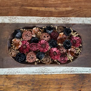 This shows a long wood dough bowl full of dried botanical berries and wood flowers. The bowl sits on a table runner on top of a coffee table.