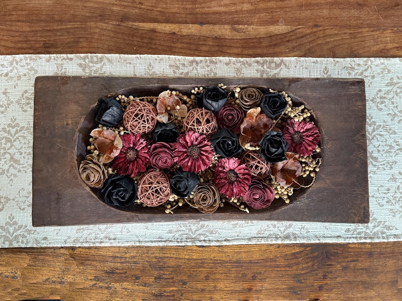 This shows a long wood dough bowl full of dried botanical berries and wood flowers. The bowl sits on a table runner on top of a coffee table.