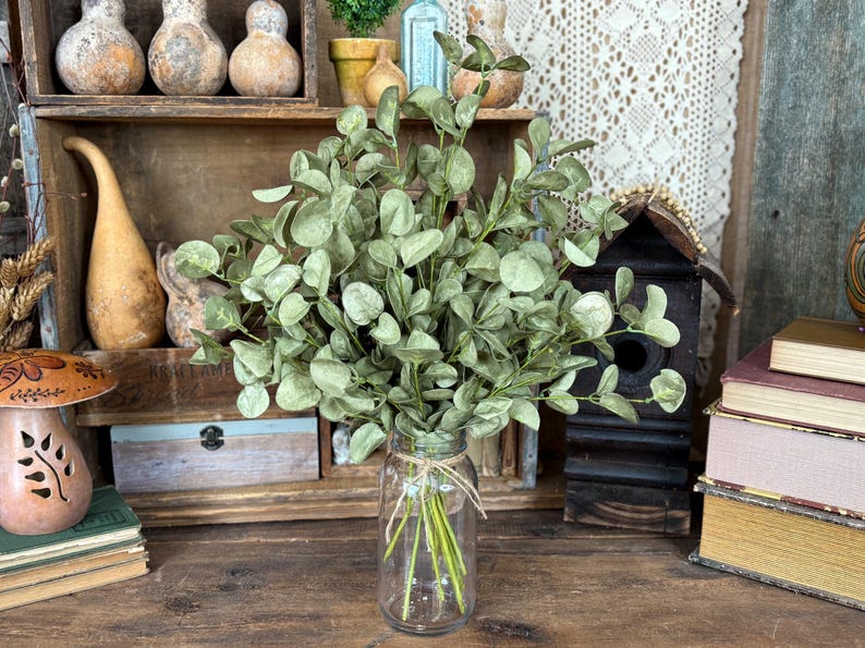 This shows a rustic farmhouse table display featuring a bouquet of green eucalyptus. The fake sprays are realistic looking arranged in a clear glass jar used as a vase. In the back is a wood bird house, stacked vintage books, and mushroom gourds.