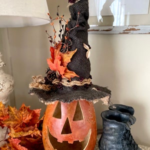 A Large handmade pumpkin witch used for fall decorating. Made from a real dried gourd. The pumpkin, named Hocus Pocus, has a tall burlap crinkled witch hat with a fall leaf arrangement. The pumpkin includes a battery-operated candle.