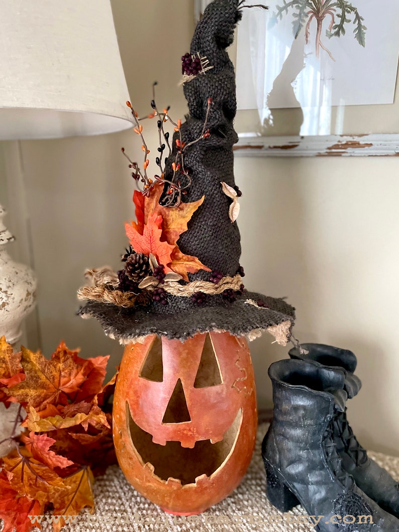 A Large handmade pumpkin witch used for fall decorating. Made from a real dried gourd. The pumpkin, named Hocus Pocus, has a tall burlap crinkled witch hat with a fall leaf arrangement. The pumpkin includes a battery-operated candle.