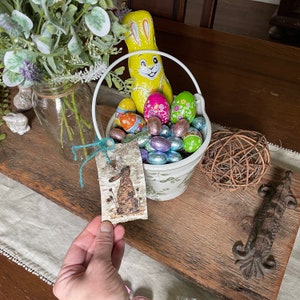 K&ouml;nnte beinhalten: Ein wei&szlig;er Keramik-Eimer gef&uuml;llt mit bunten Schoko-Ostereiern und einem gelben Schokoladenhasen. Ein kleines Holzschild mit einer braunen Hasenillustration ist am Eimer befestigt.