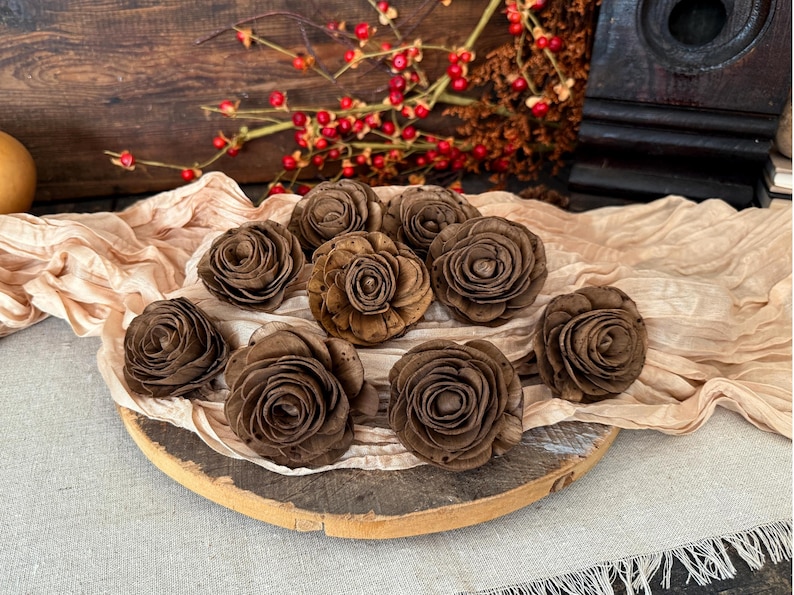 This shows several wood flowers that look like ranunculus, in a shade of chocolate brown. They are laid our on a beige table runner, on top a a vintage style bread board. Bittersweet berries and a dark wood bird house are in the background.