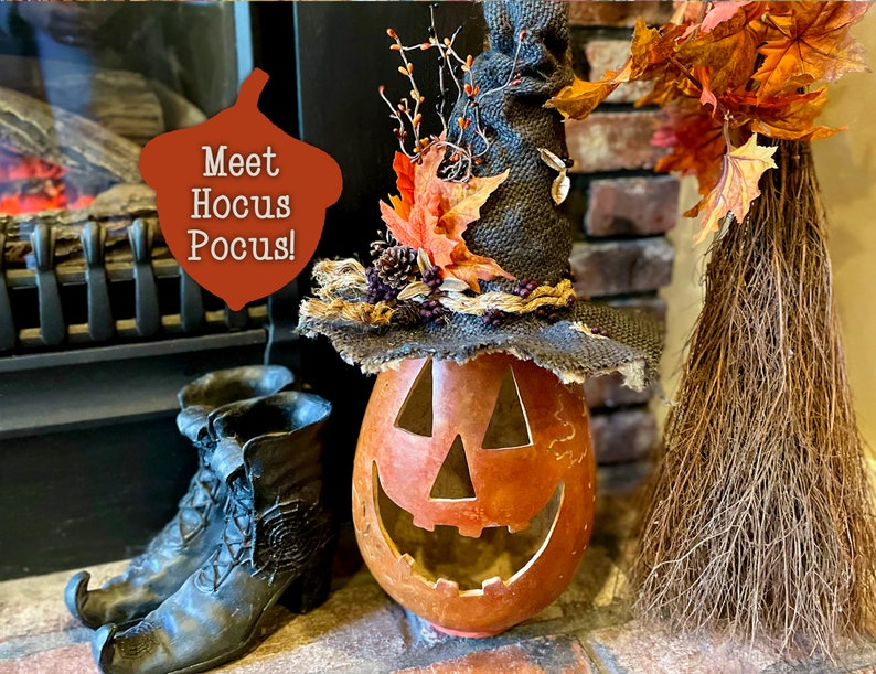 A Large handmade pumpkin witch used for fall decorating. Made from a real dried gourd. The pumpkin, named Hocus Pocus, has a tall burlap crinkled witch hat with a fall leaf arrangement. The pumpkin includes a battery-operated candle.