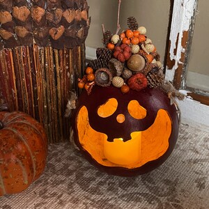 This is a handmade pumpkin decoration. A battery-operated candle lights the pumpkin. It is a real dried gourd, with a carved ghostly facial expression. The shell has been stained a burgundy color. There is a fall berry arrangement on the top.