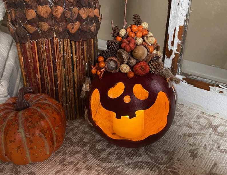 This is a handmade pumpkin decoration. A battery-operated candle lights the pumpkin. It is a real dried gourd, with a carved ghostly facial expression. The shell has been stained a burgundy color. There is a fall berry arrangement on the top.