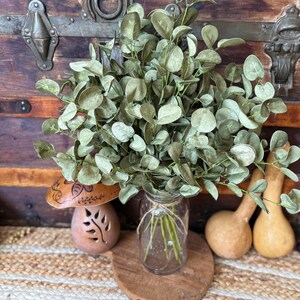 This shows a rustic greenery arrangement  featuring a bouquet of green eucalyptus. The fake sprays are realistic looking arranged in a clear glass jar used as a vase. In the back is antique trunk and dried gourds.