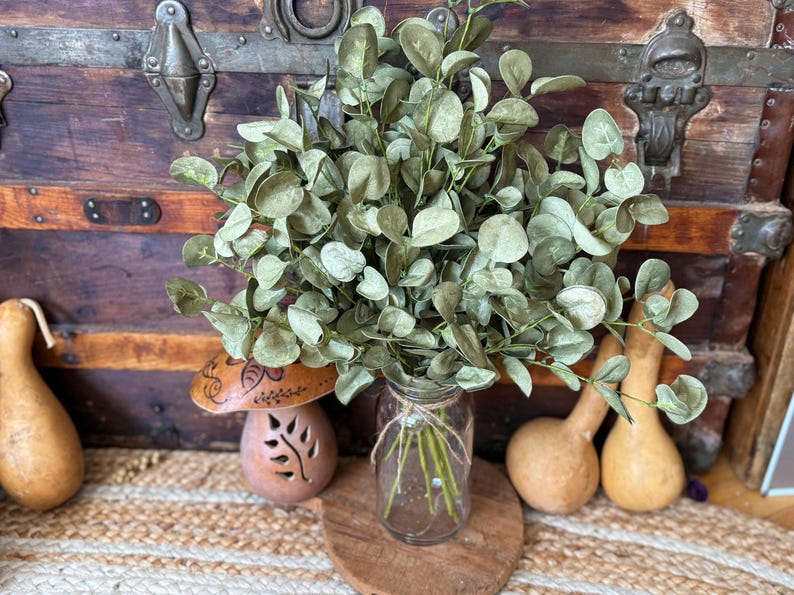 This shows a rustic greenery arrangement  featuring a bouquet of green eucalyptus. The fake sprays are realistic looking arranged in a clear glass jar used as a vase. In the back is antique trunk and dried gourds.