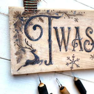 Pode incluir: Uma placa de madeira com as palavras "Twas the" gravadas nela. A placa &eacute; decorada com uma rena, azevinho e flocos de neve. A placa est&aacute; pendurada em um clipe de metal enferrujado.