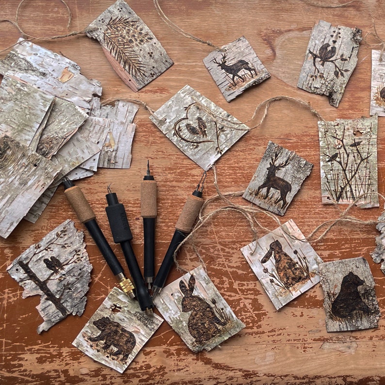This image shows a variety of handmade birch bark tags. They are laid out on an antique trunk. Pyrography tools lay next to them. The bark is torn into 3-inch pieces. Each tag has a different wood animal burned onto it.