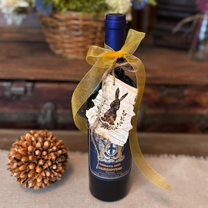 This image shows a handmadegift tag tied to a wine bottle gift. The tag is made from real birch bark. I  have wood-burned a bunny rabbit onto the tag. A yellow ribbon is tied to the neck of the wine bottle. A decorative rattan ball sits to the left.