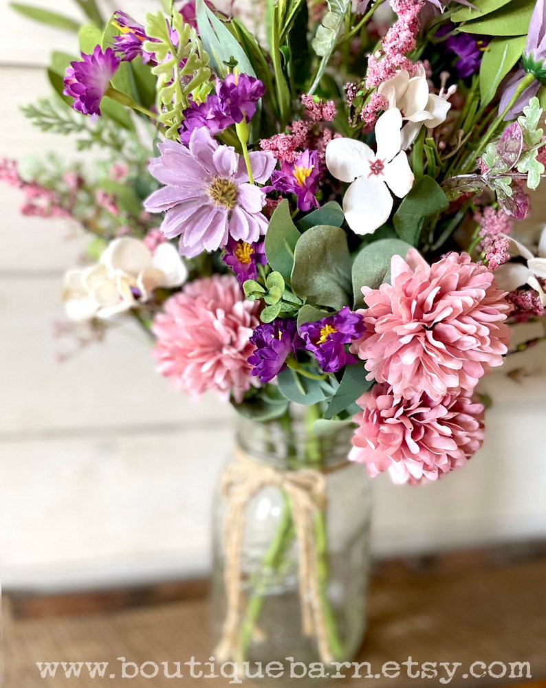 This image shows an artificial bouquet of flowers in a mason jar vase. It features purple daisies, pink mums, and white hydrangeas.