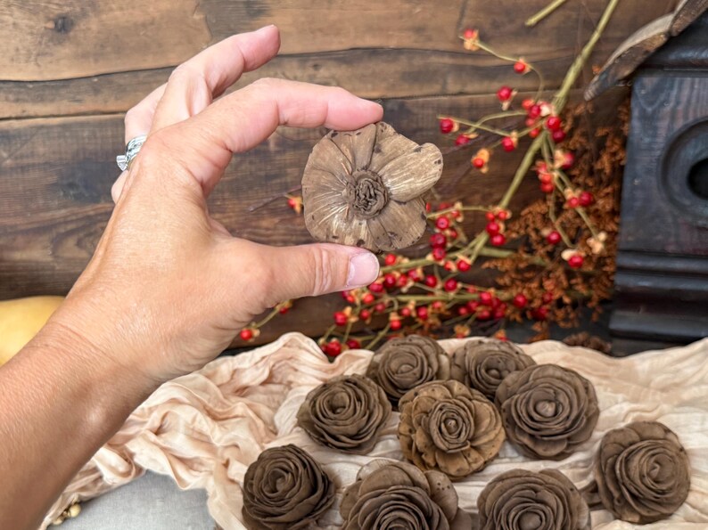 This shows me holding the wood flower blossom so you can see the back view. Several wood flowers are laid out in the background on a decorative table scarf. You can also see bittersweet branches in the back.