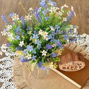 Könnte beinhalten: Ein Blumenarrangement in einer durchsichtigen Glasvase mit blauen, weißen und lila Blumen. Die Vase steht auf einer Jutematte, mit einem Buch und einem Holzblock mit der Aufschrift "choose HAPPY". Der Hintergrund ist eine Holzoberfläche.