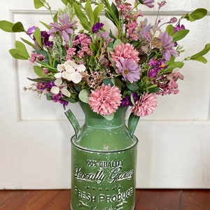 This image shows an artificial bouquet of flowers in a farmhouse green milk can. It features purple daisies, pink mums, and white hydrangeas.