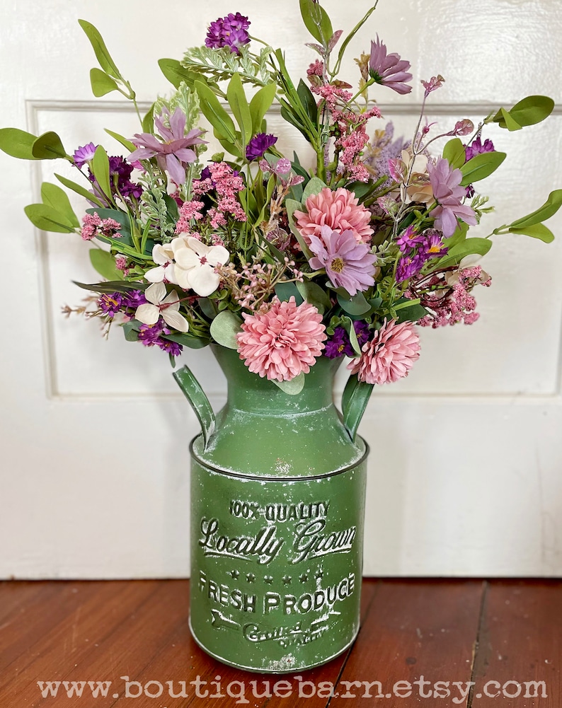 This image shows an artificial bouquet of flowers in a farmhouse green milk can. It features purple daisies, pink mums, and white hydrangeas.