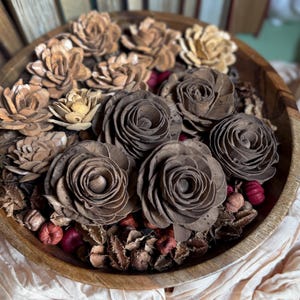 This shows several wood flower blossoms, arranged in a wooden bowl with other dried botanicals,  sitting on an accent table. Several vintage books are standing in the background.