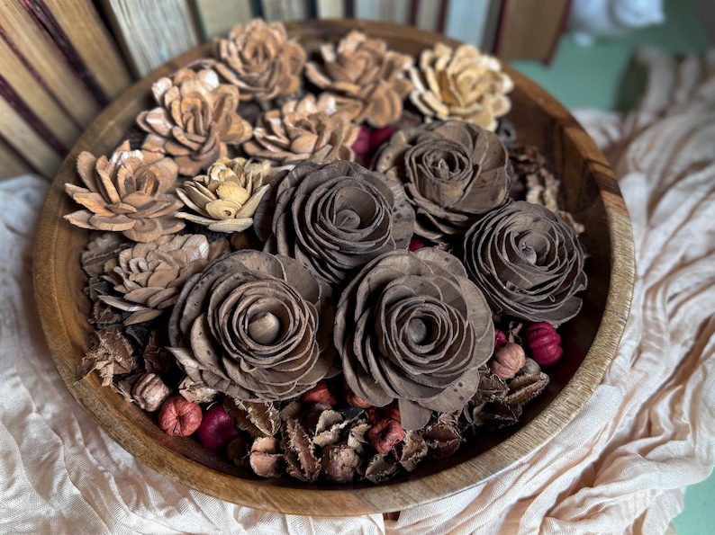 This shows several wood flower blossoms, arranged in a wooden bowl with other dried botanicals,  sitting on an accent table. Several vintage books are standing in the background.