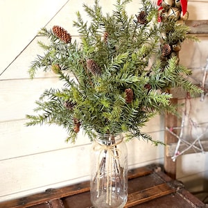 May include: A bouquet of artificial pine sprigs with brown pine cones in a clear glass jar with a twine bow.