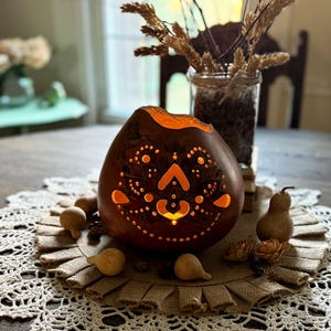 May include: A brown gourd-shaped candle holder with detailed cut-out patterns, glowing with an orange light. It's surrounded by decorative gourds and rests on a burlap and lace doily, creating a rustic, autumnal theme.