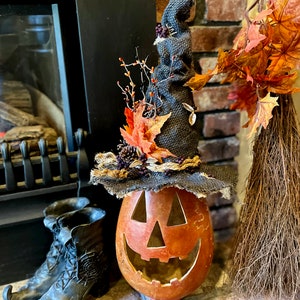 A Large handmade pumpkin witch used for fall decorating. Made from a real dried gourd. The pumpkin, named Hocus Pocus, has a tall burlap crinkled witch hat with a fall leaf arrangement. The pumpkin includes a battery-operated candle.