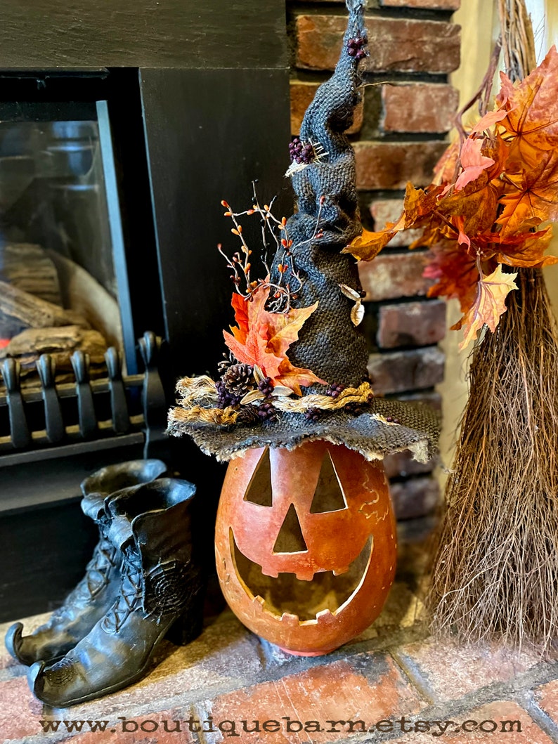 A Large handmade pumpkin witch used for fall decorating. Made from a real dried gourd. The pumpkin, named Hocus Pocus, has a tall burlap crinkled witch hat with a fall leaf arrangement. The pumpkin includes a battery-operated candle.