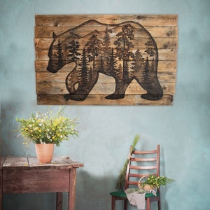 This is a large wood wall hanging featuring a bear and a forest scene. The artwork is wood burned by hand onto a pallet of reclaimed fence. The wall decor is hanging over a farmhouse table on a blue wall.