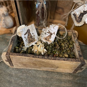 This shows a wooden bowl filler display. It has my handmade birch bark tags nestled in a wooden box of dried green botanicals. A decorative bundle of books has a fox tag tied to it.